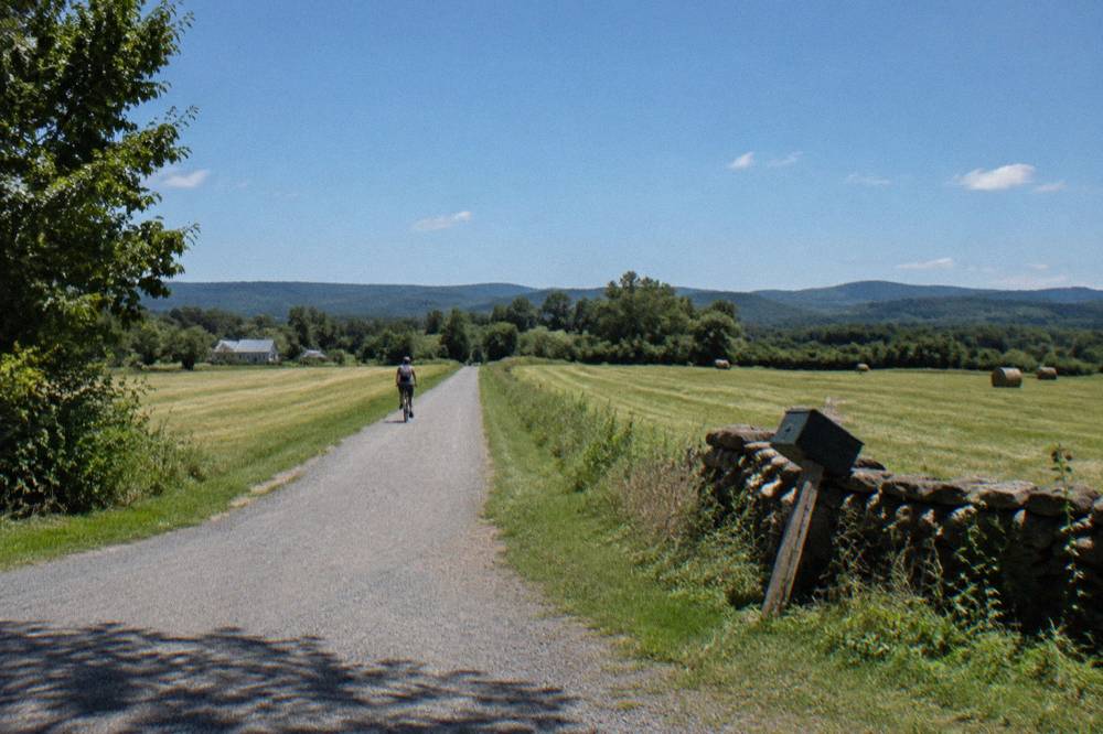 A long straight rail trail crossing open farmland with mountains on the horizon under a blue sky