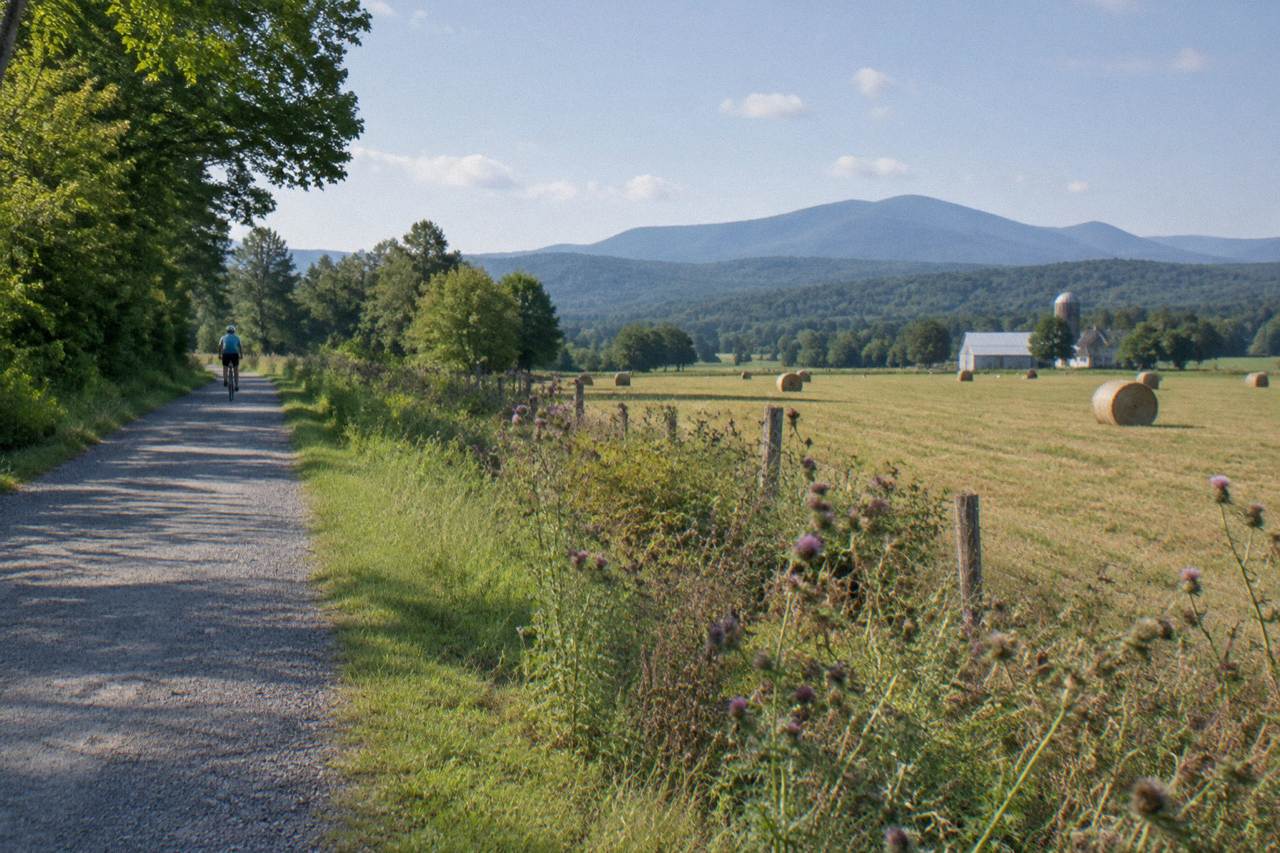 A wide crushed stone rail trail curving through green Vermont farmland with wooded hills behind