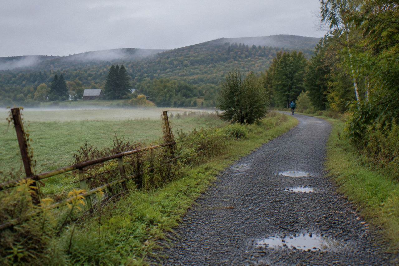 A crushed stone rail trail curving through green Vermont countryside with wooded hills behind