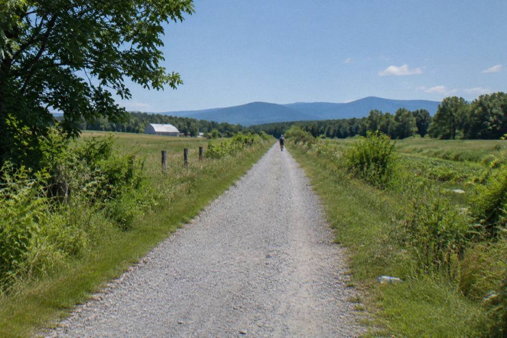 A long straight rail trail crossing open farmland with mountains on the horizon