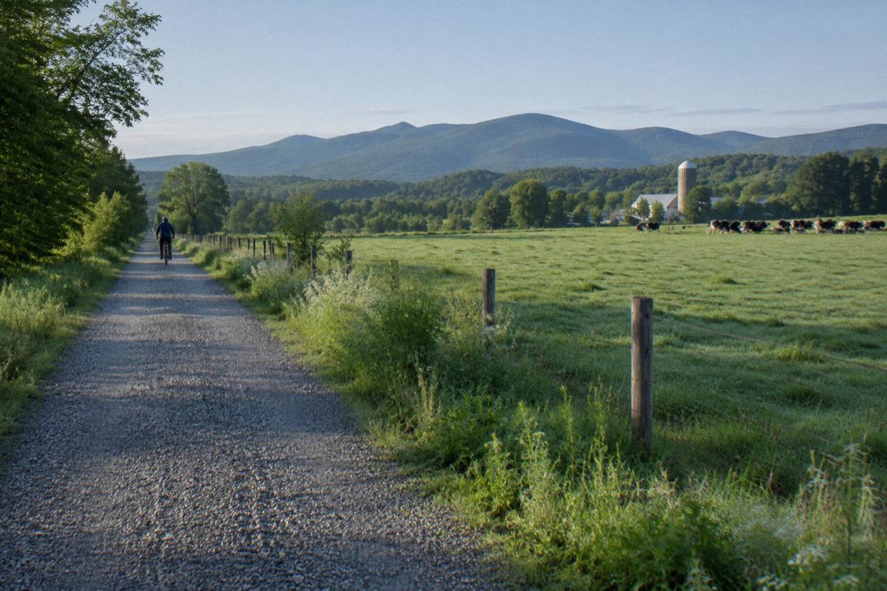 A crushed stone rail trail stretching through green Vermont farmland with wooded hills behind