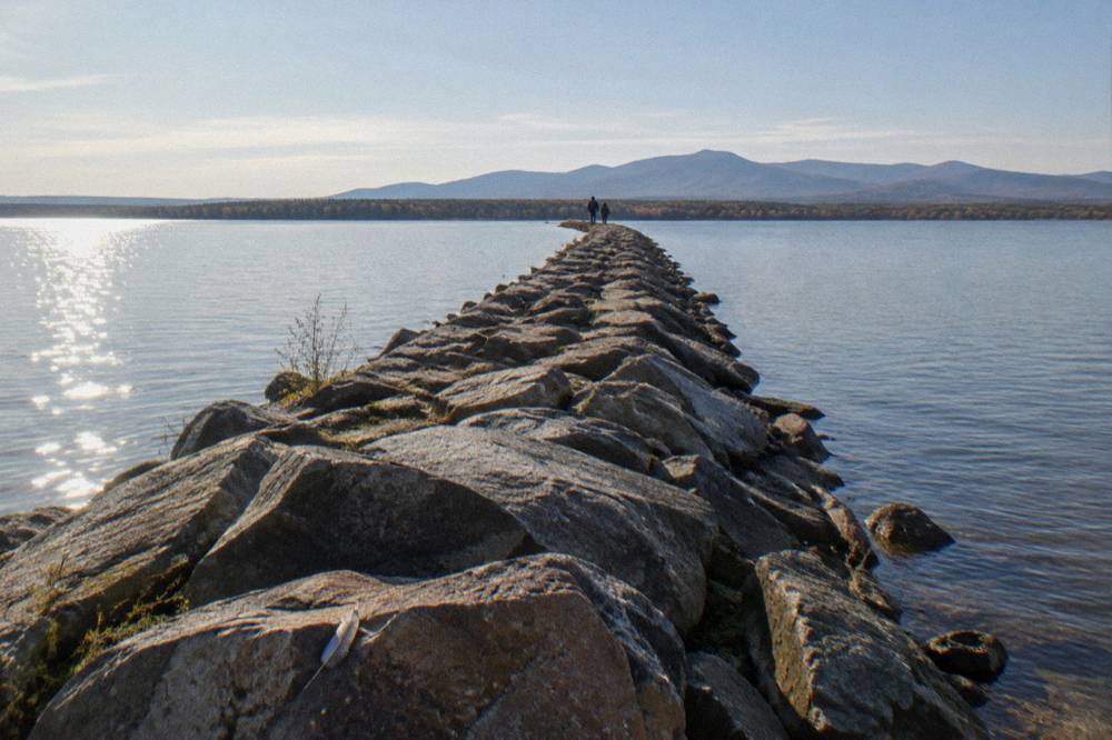 A gravel causeway path stretching straight across a wide lake with distant mountains on the horizon