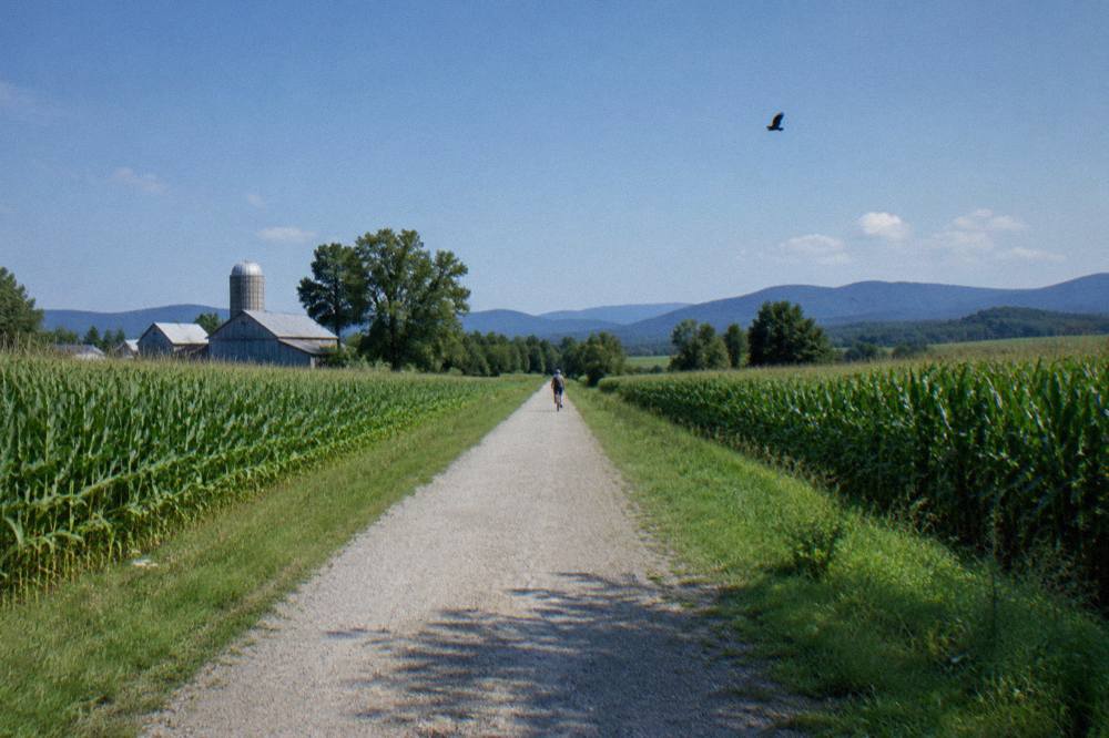 A wide crushed stone rail trail running straight through green farmland with hills behind