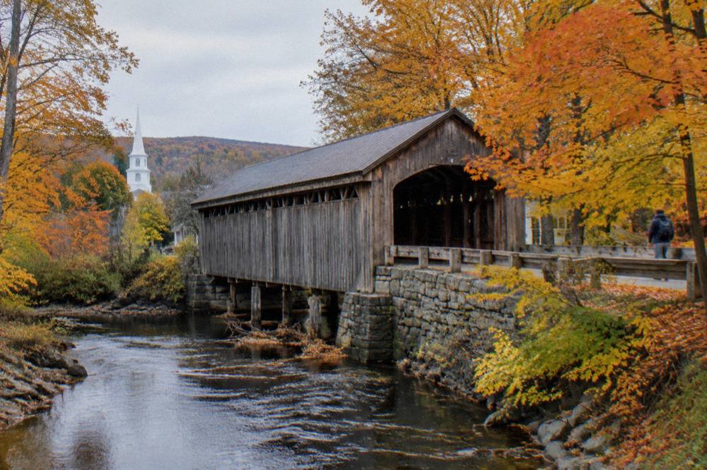 A covered bridge over a stream lined with orange and yellow trees in a Vermont village