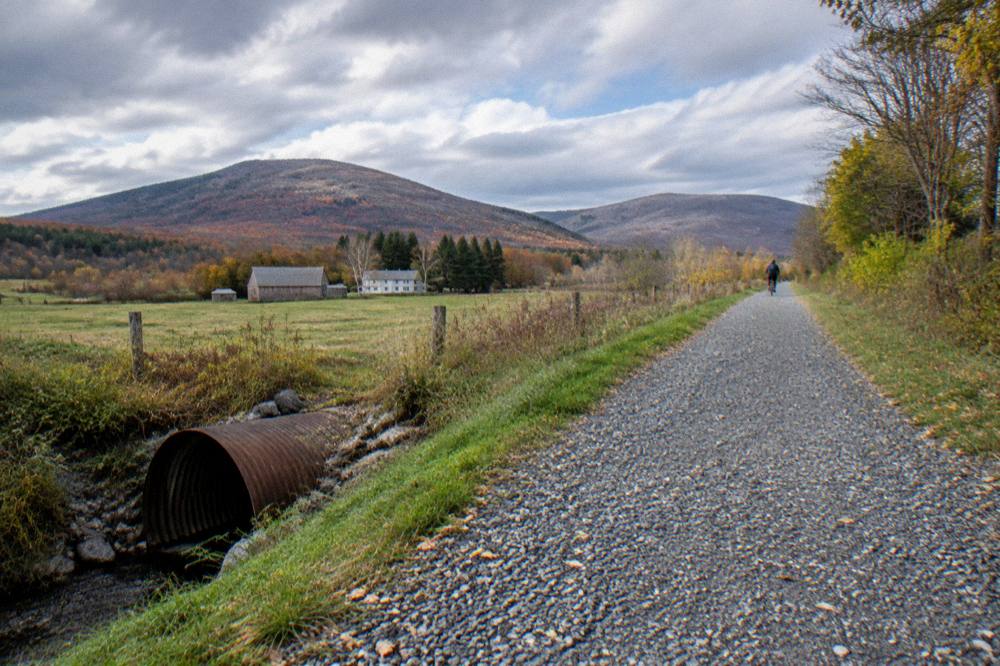 A wide crushed stone rail trail crossing open farmland with hills in fall color beyond