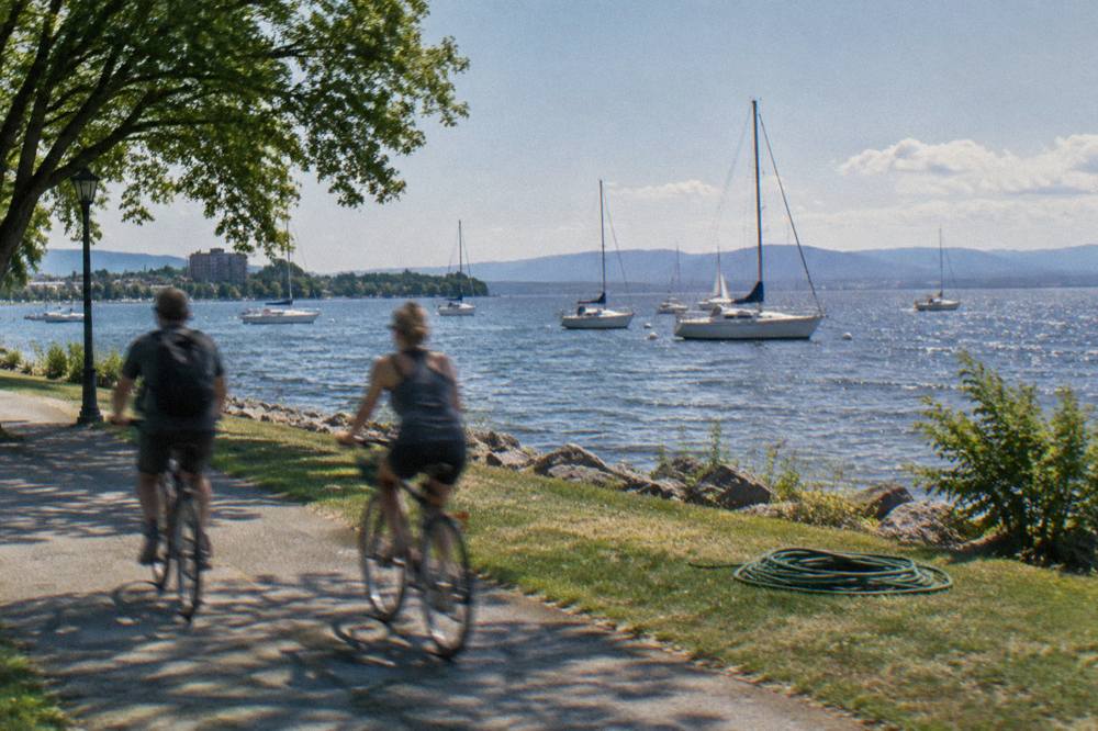 Cyclists riding a paved path beside a lake with sailboats moored offshore