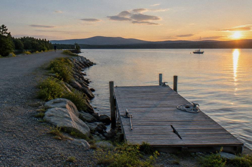 A wooden pontoon dock extending from a gravel causeway into calm lake water