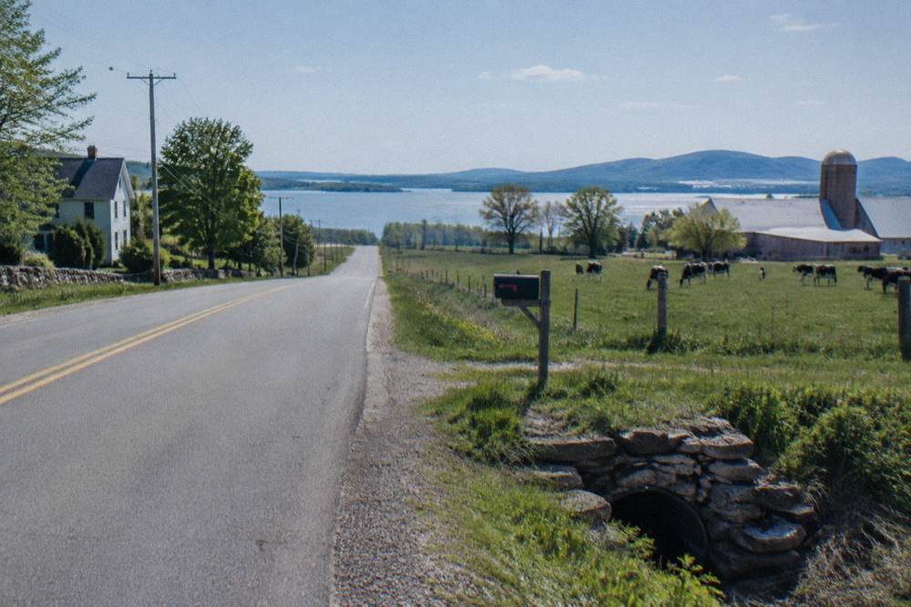 A quiet rural road passing farm fields with lake water visible between distant hills