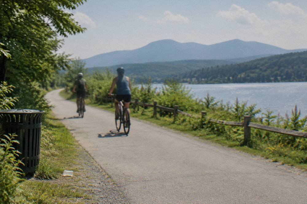 Cyclists riding a paved path beside a lake with mountains on the horizon