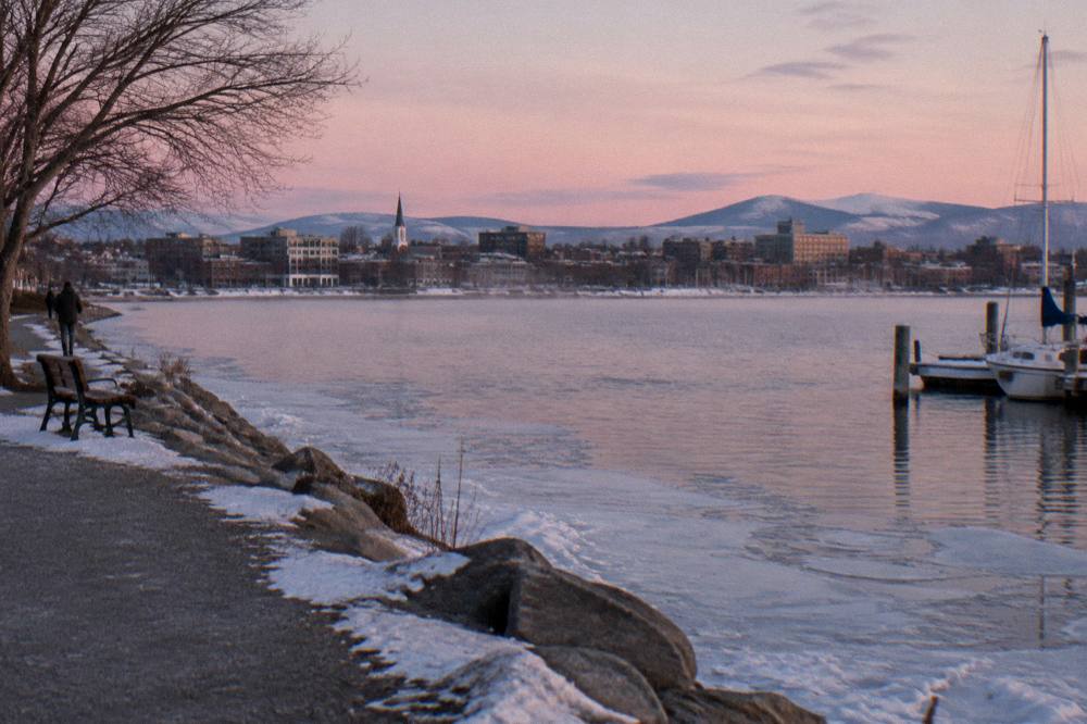 A small city skyline seen across a lake at dusk from a shoreline path