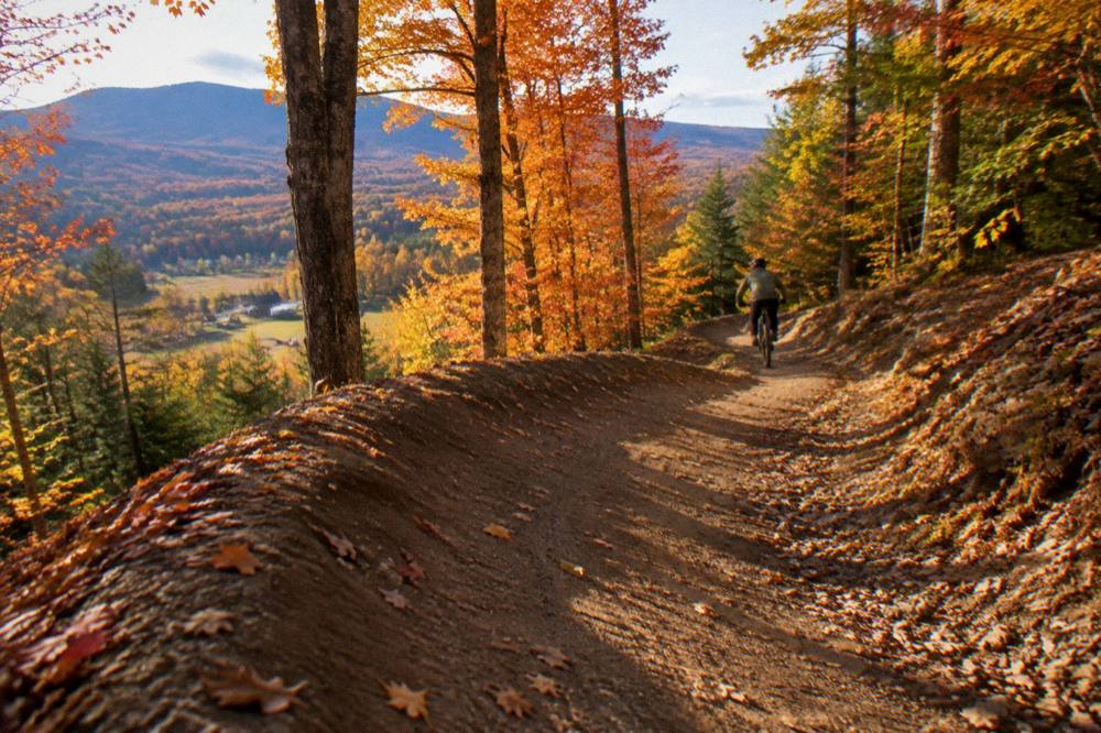 A mountain biker rolling through bermed singletrack beside tall maples in fall color