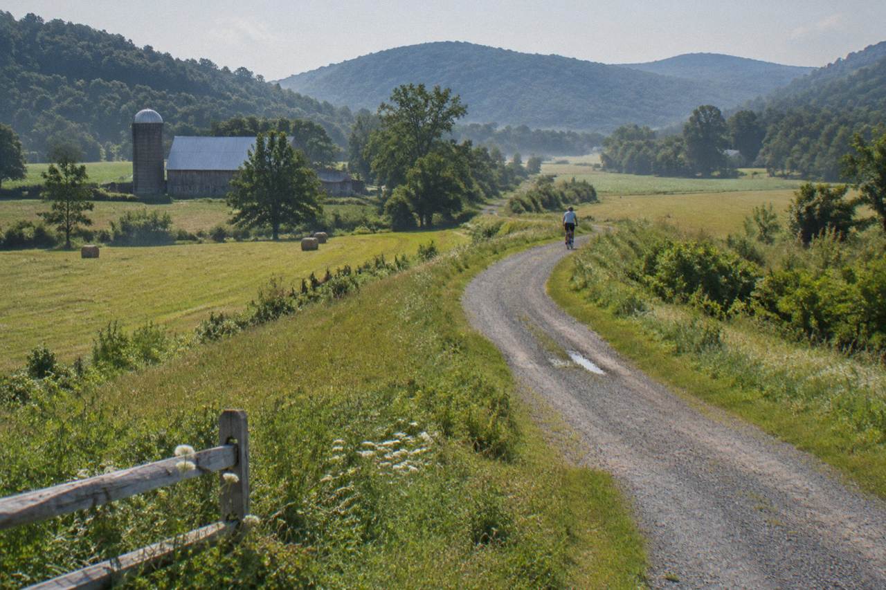 A crushed stone rail trail curving through green Vermont farmland with wooded hills behind