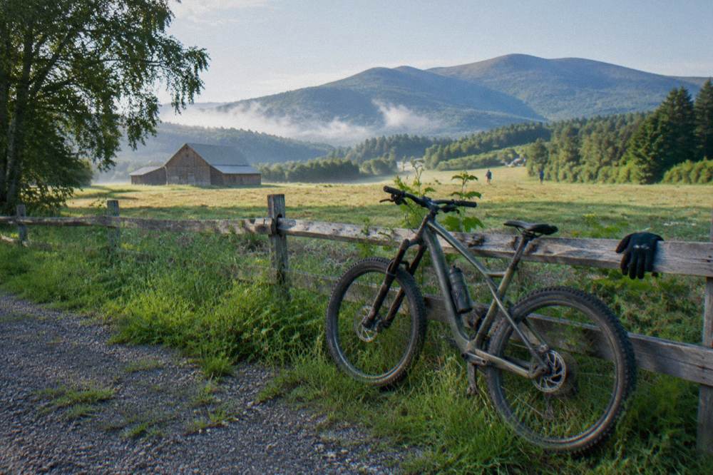 A mountain bike leaning against a wooden fence beside a Vermont farm field trailhead