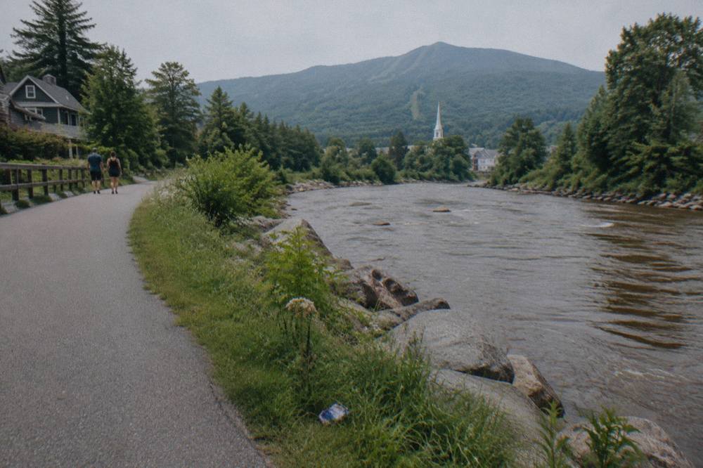 A paved recreation path winding beside a rocky river with forested hills behind it