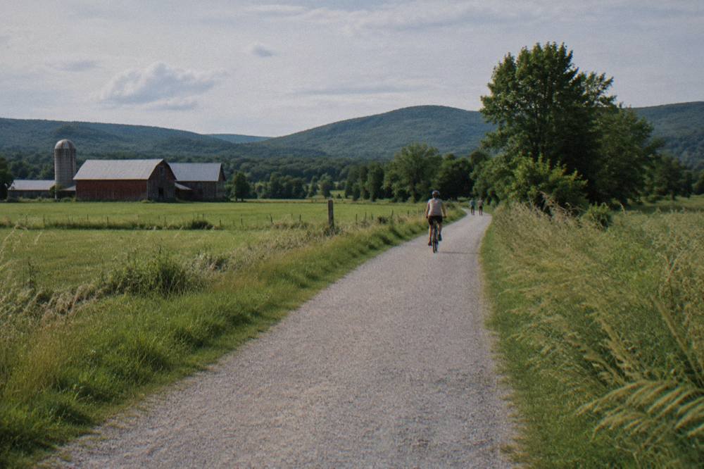 A wide crushed stone rail trail curving through green farm fields with hills beyond