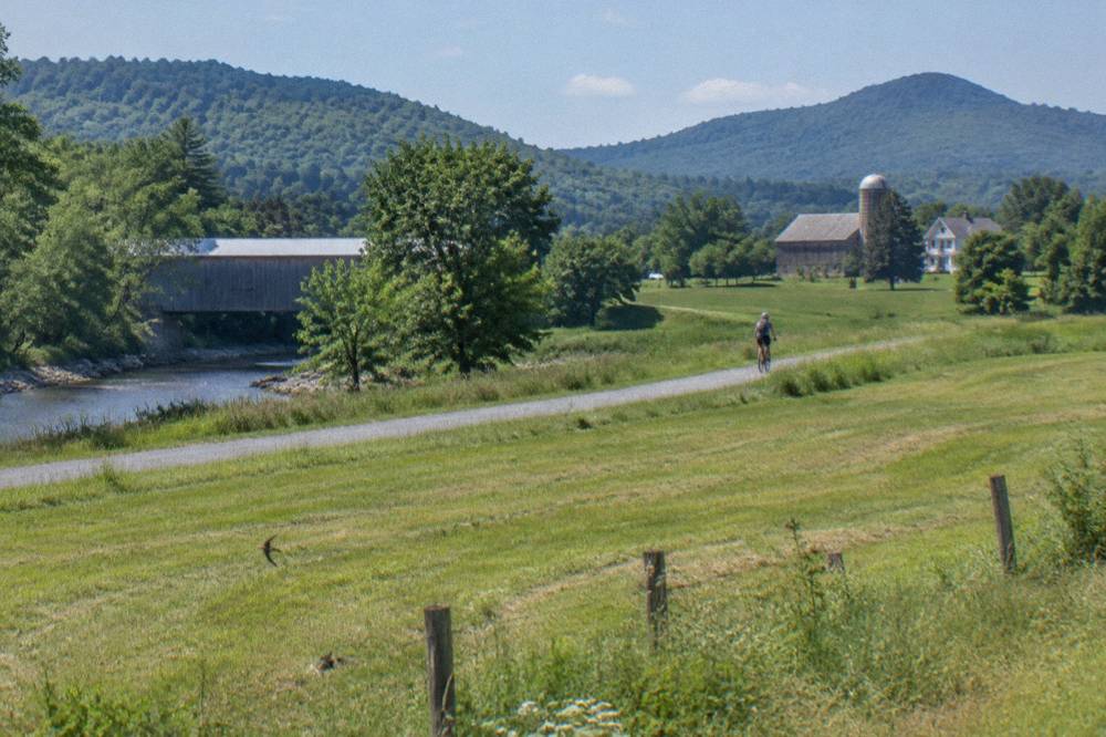 A cyclist riding a rail trail of packed crushed stone through green Vermont farmland