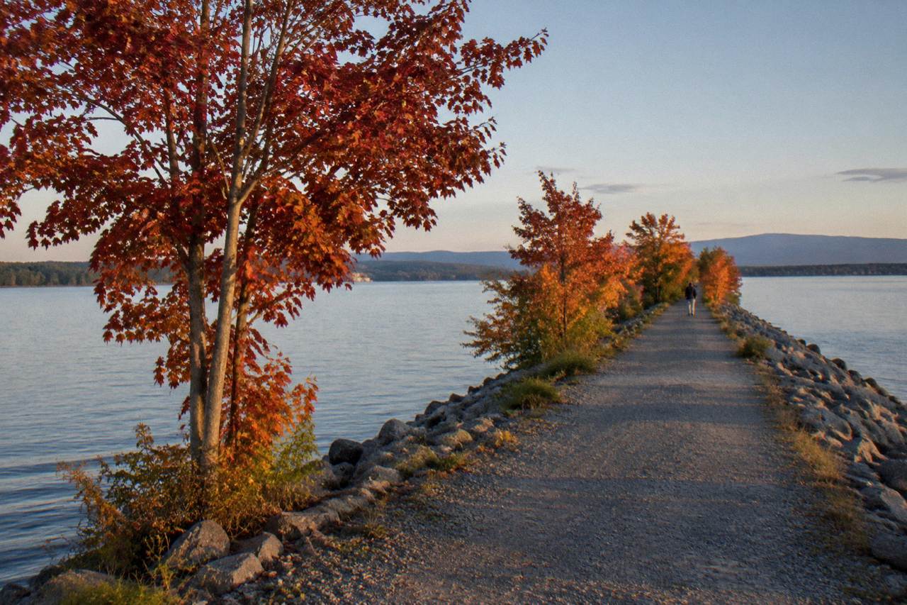 A paved bike path lined with red and orange maple trees beside a quiet Vermont valley