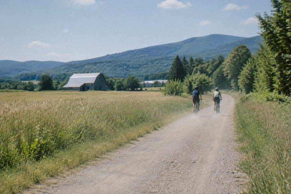 Two cyclists riding side by side on a wide dirt road between Vermont hay fields