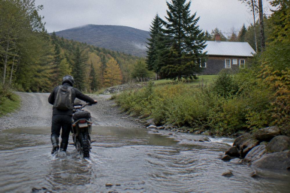 A rider walking a loaded gravel bike across a shallow stream ford on a forest road