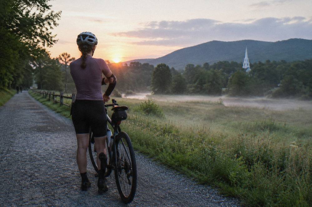 A cyclist adjusting arm warmers beside a bicycle on a tree-lined rural road in morning light