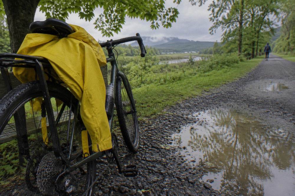 A rain jacket strapped over a bicycle seat bag on a parked bike beside a wet gravel trail