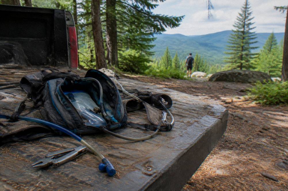A hydration pack and trail tools open on a tailgate beside a mountain bike in a forest clearing